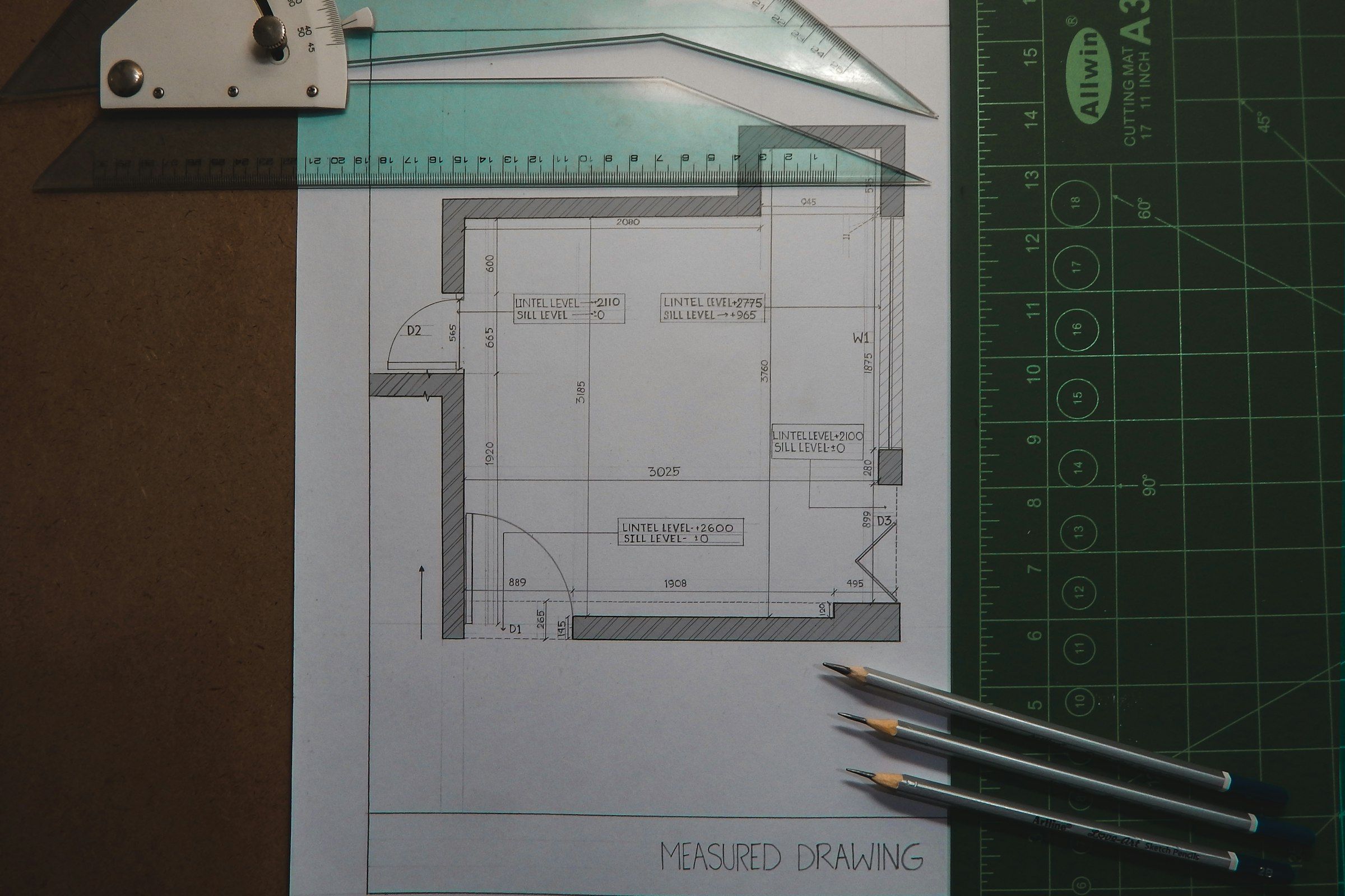 A hand-drafted measured floor plan on a cutting mat, with a drafting square and pencils