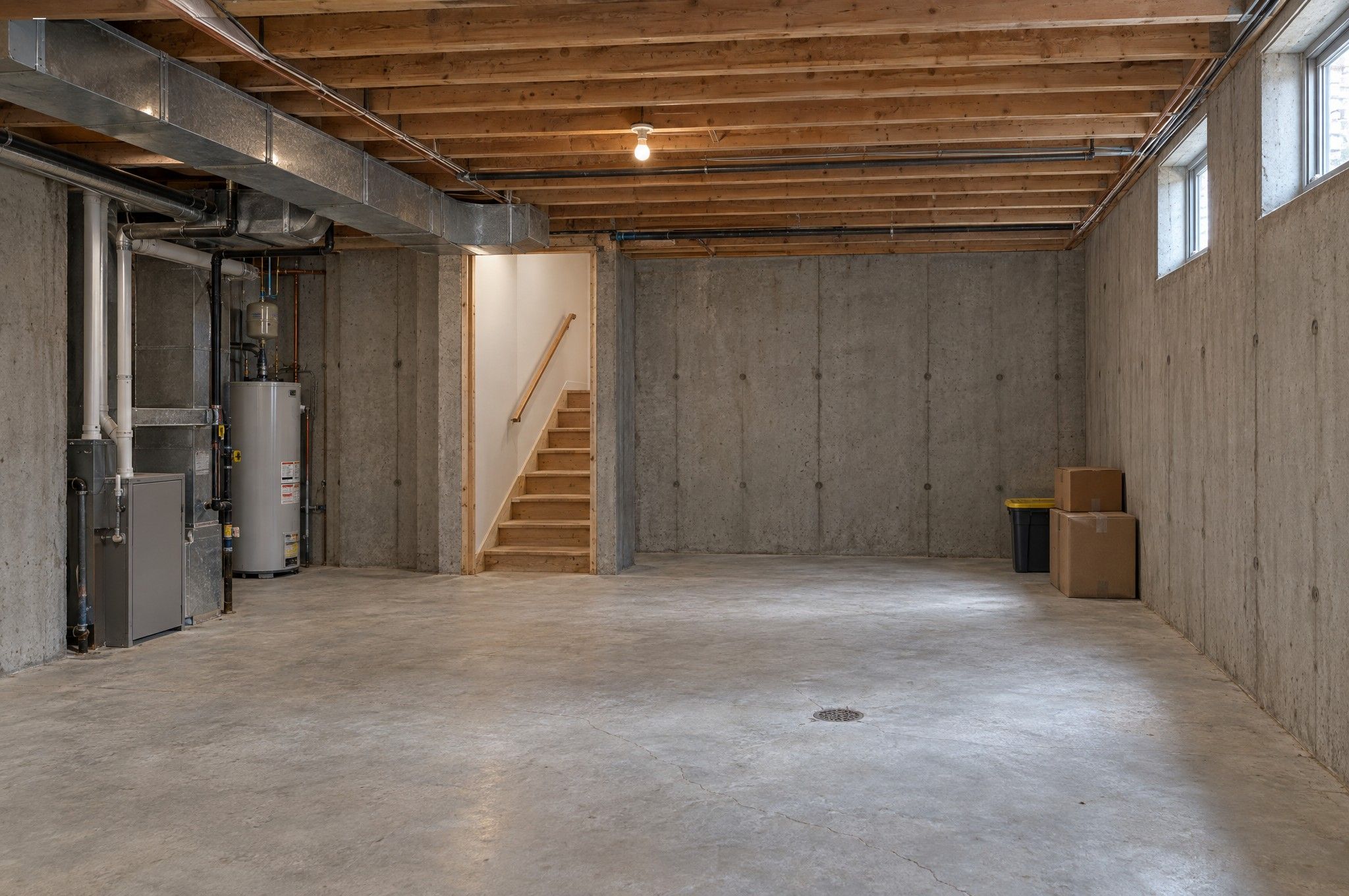 Unfinished basement: exposed joists and ductwork, bare concrete walls and slab, furnace and water heater, raw wood stair