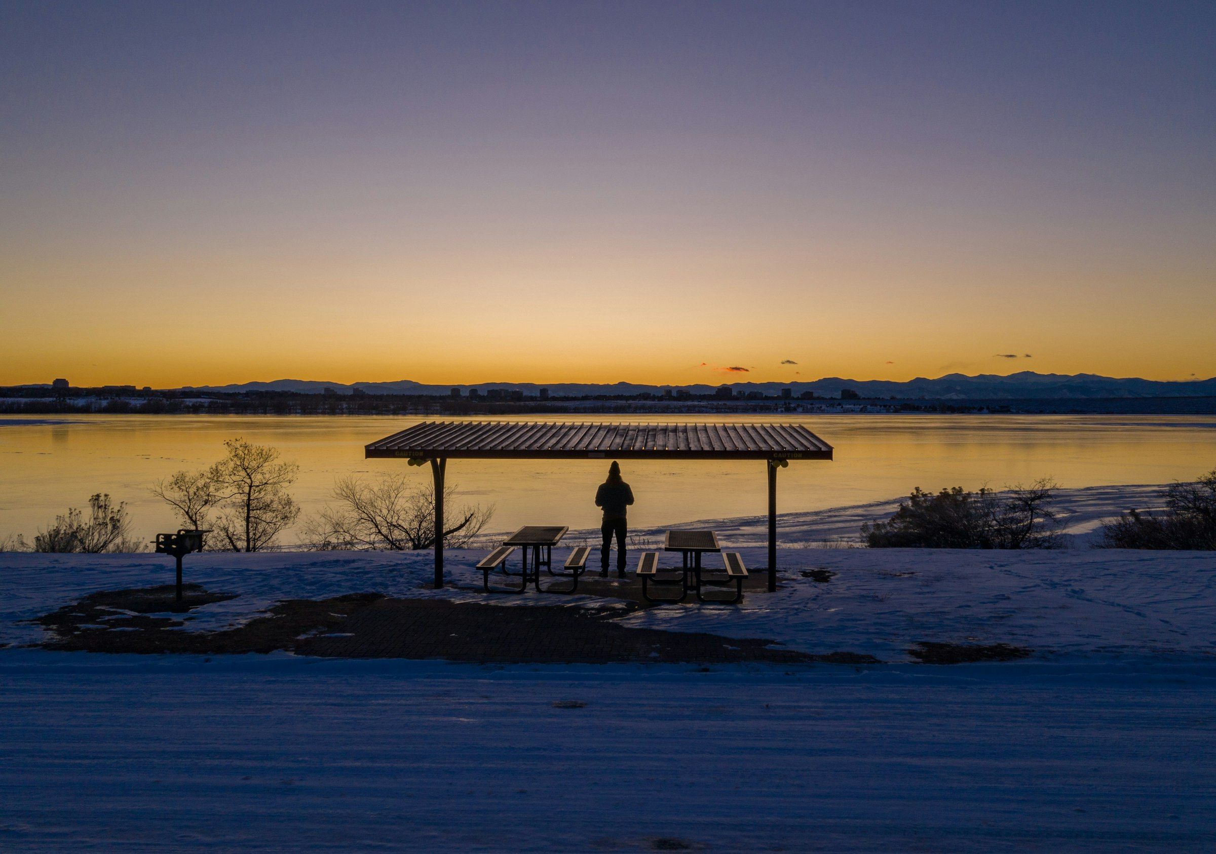 A person standing under a picnic shelter beside a reservoir in Aurora at sunset