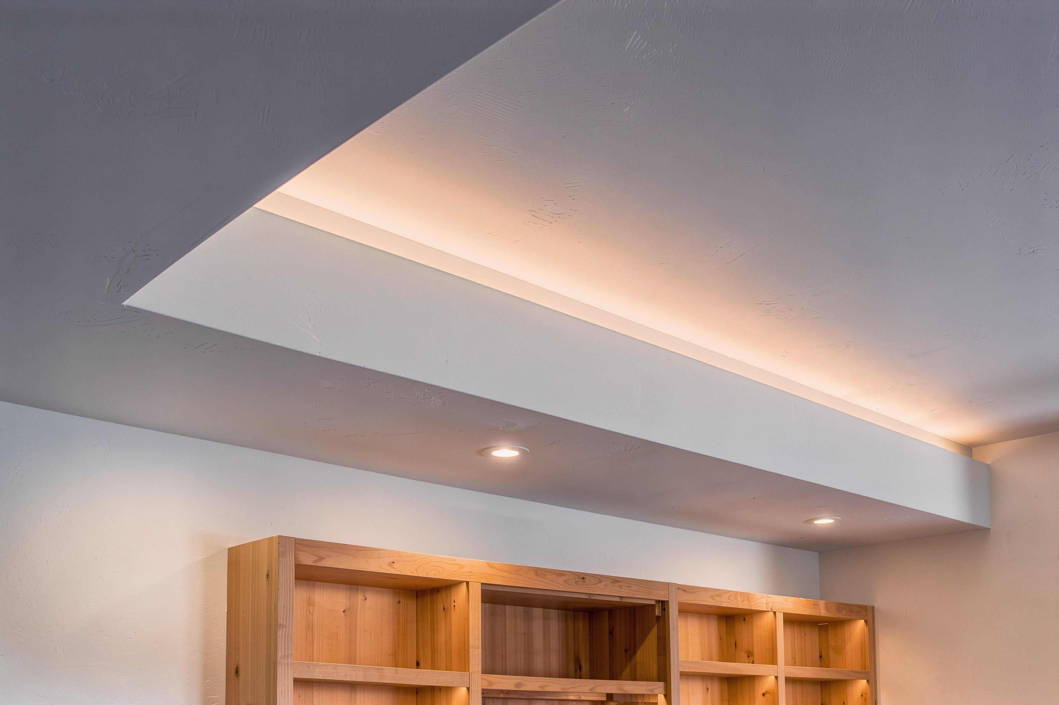 A painted drywall soffit along a basement ceiling, with warm LED cove light washing the ceiling from a hidden ledge, above white-oak shelving