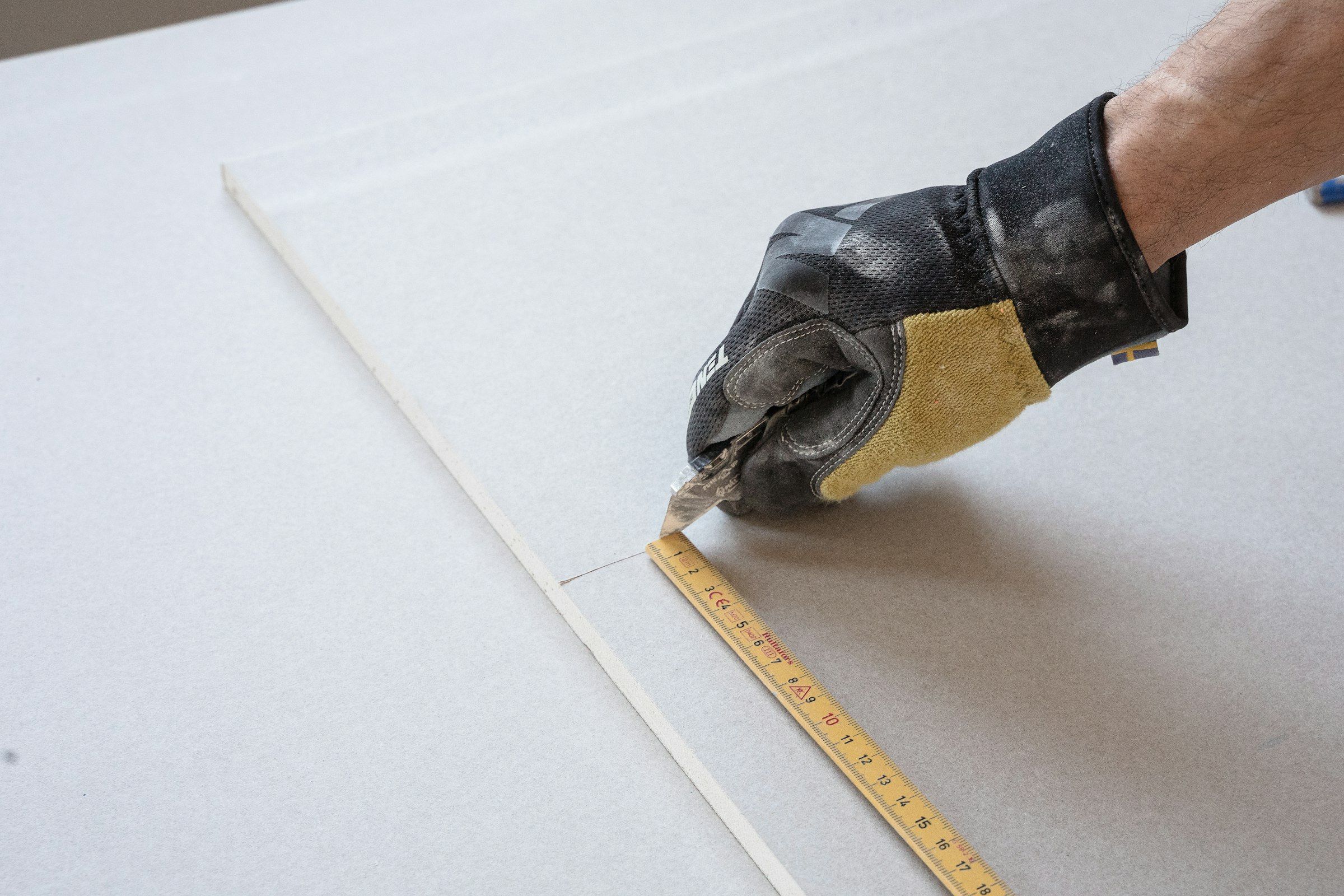 A gloved hand cutting drywall along a folding rule